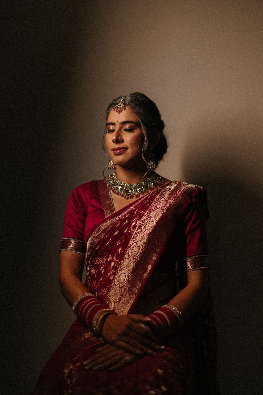Indian bride in a deep red silk saree with gold jewelry and maang tikka in a moody portrait.