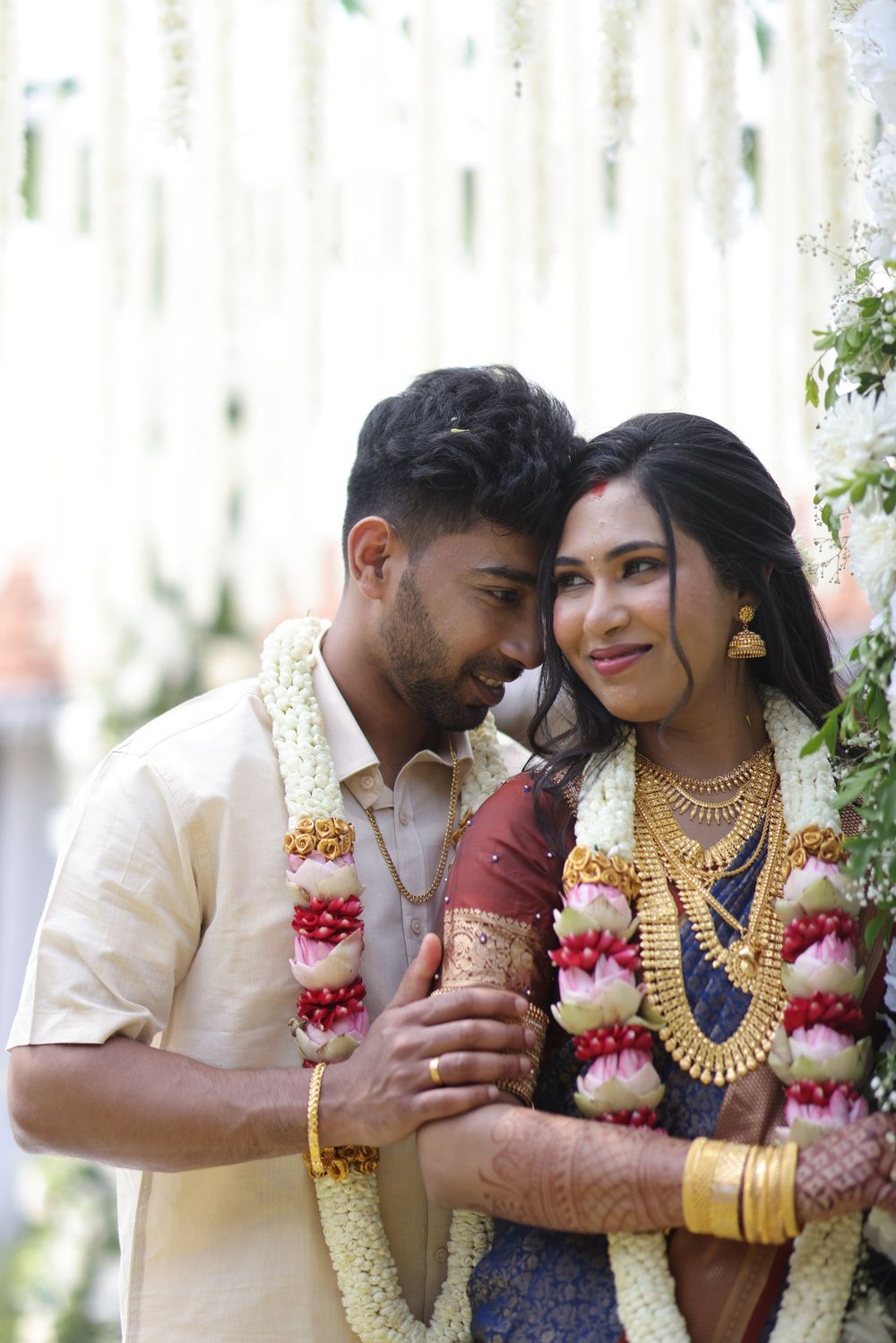Indian couple in traditional wedding attire with floral garlands posing together at a decorated ceremony venue.