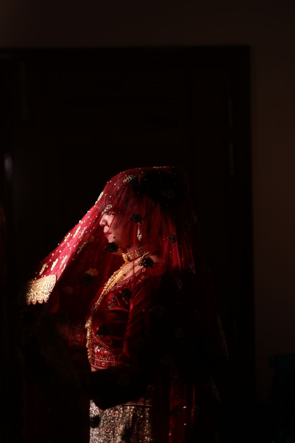 A bride in a red bridal lehenga and dupatta stands elegantly in dramatic lighting.