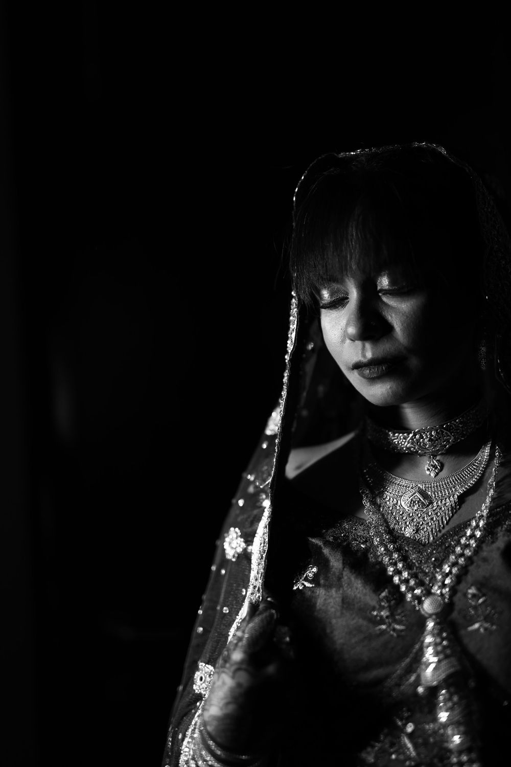 Black and white portrait of a woman in traditional South Asian bridal attire with veil and jewelry, eyes downcast.