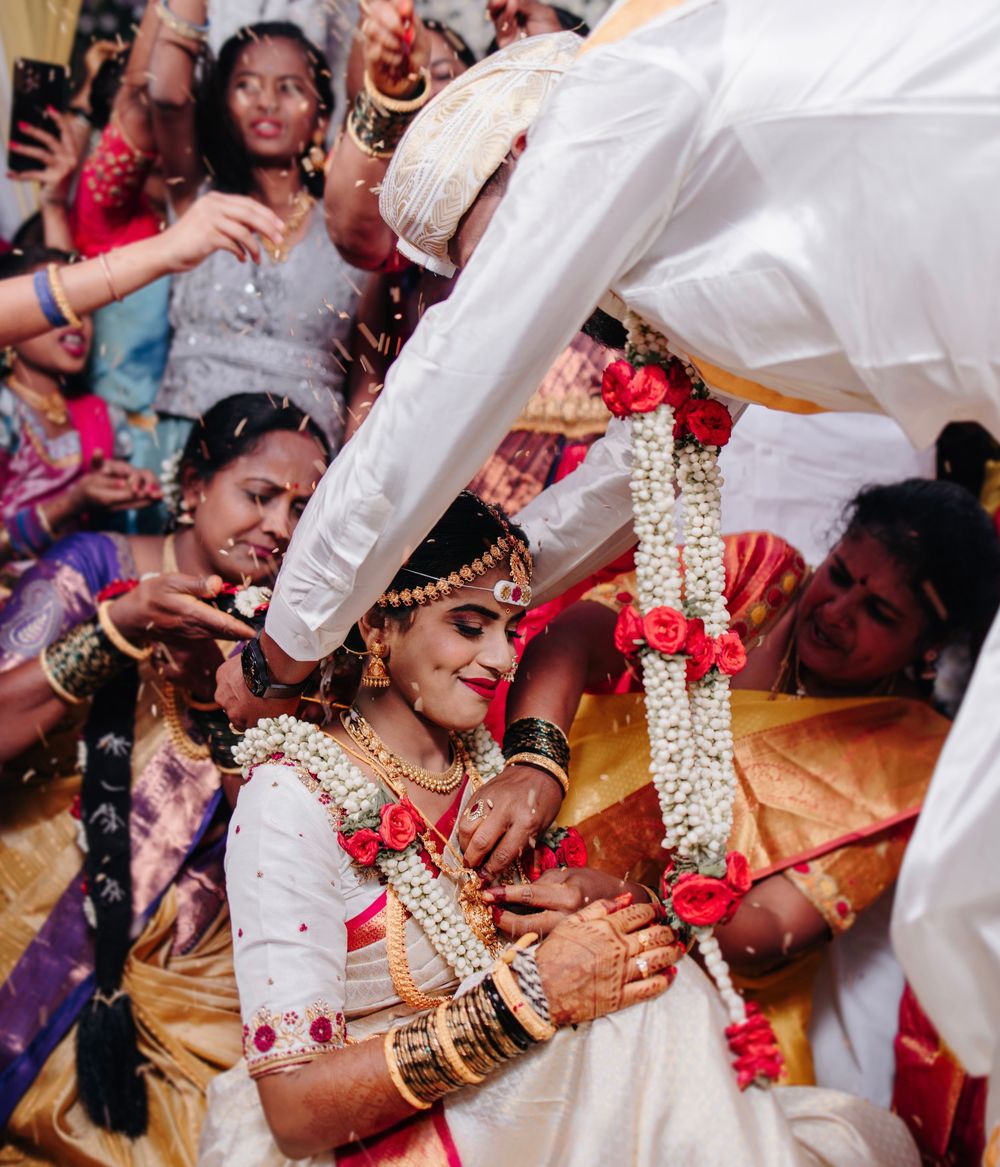 Bride smiling during South Indian wedding ceremony as groom places garland amid celebrating guests.