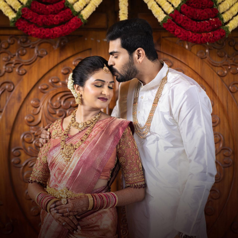 A couple in traditional South Indian wedding attire, bride in pink saree with gold jewelry, groom in white kurta.