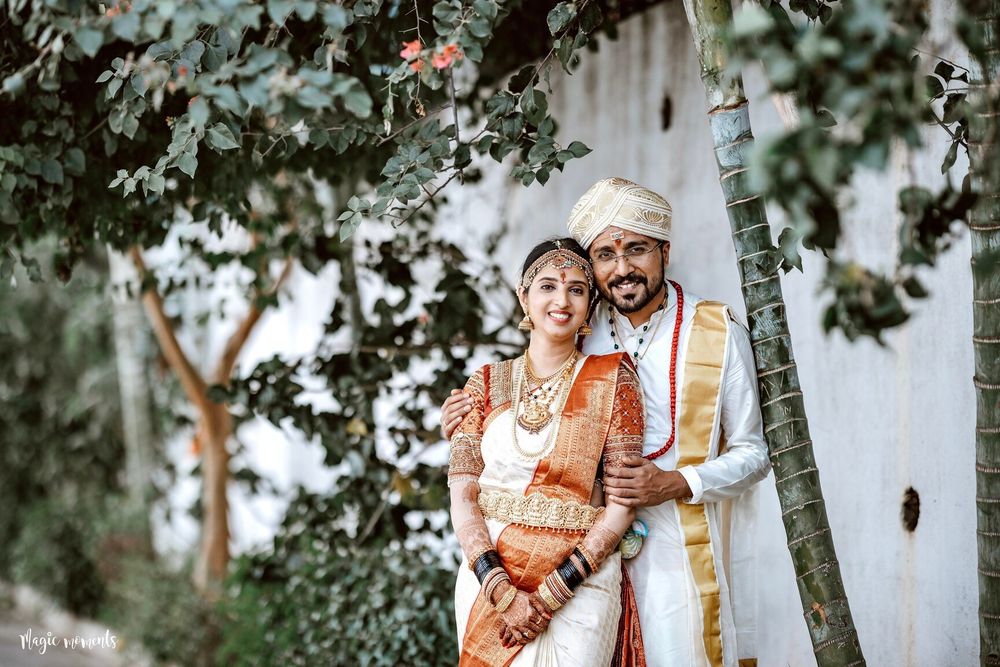 Indian couple in traditional wedding attire posing outdoors under green foliage, bride in red saree and groom in white dhoti.
