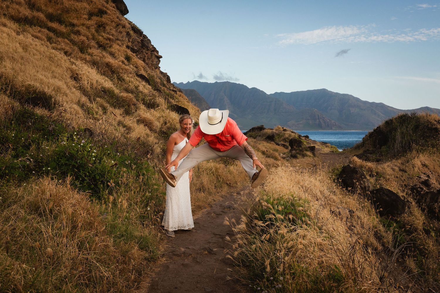 Couple enjoying a scenic Hawaii trail with golden hills and ocean views. Cowboy groom wears a coral shirt and cowboy hat.  Hawaii Wedding