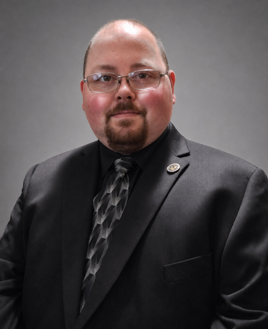 Professional headshot of a bald man with glasses and a beard wearing a black suit and patterned tie.