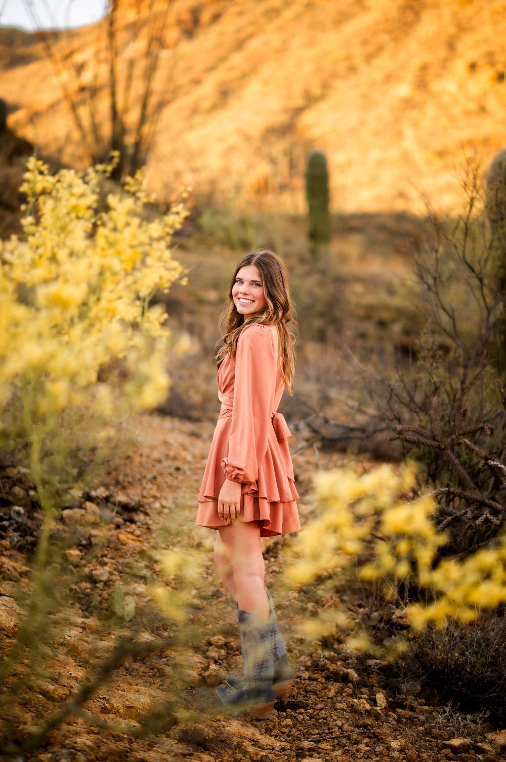 Young woman in coral dress smiling in desert landscape with yellow wildflowers and saguaro cactus.