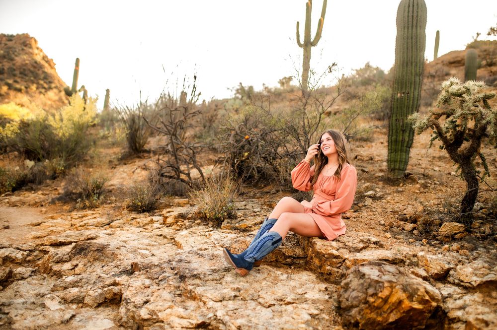 Woman in pink dress and blue boots sits on desert rocks surrounded by saguaro cacti in Arizona landscape.