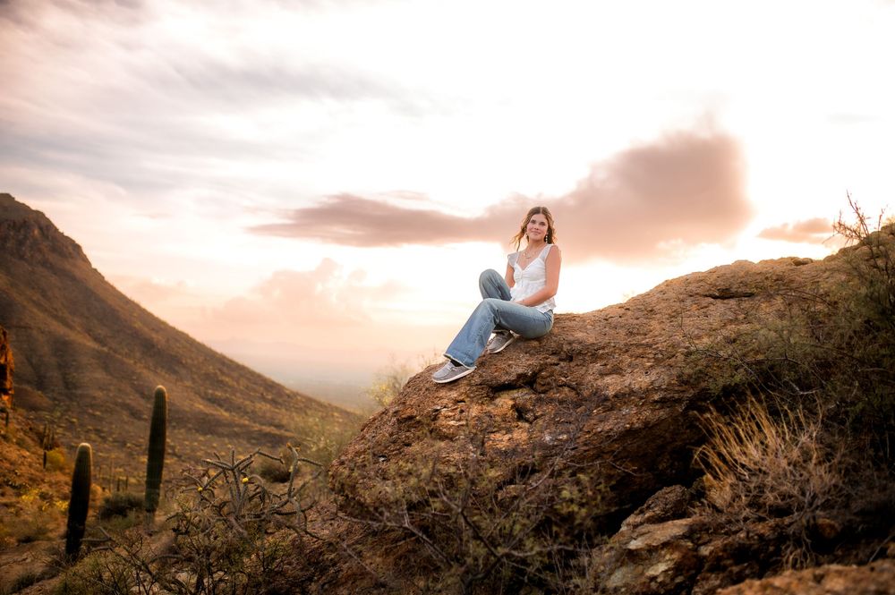 Woman sitting on a rocky desert hillside at sunset with cacti and mountain valley views in the background.