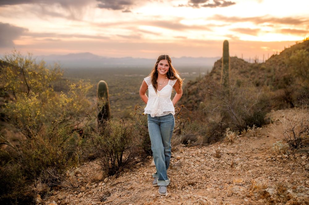 Woman in white top and jeans walks a desert trail at sunset with saguaro cacti and mountains in background.