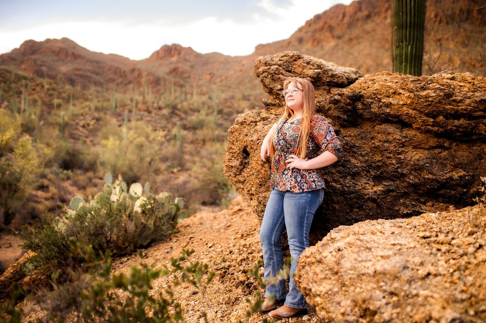 Young woman in floral top and jeans leaning on desert rocks with saguaro cacti and mountains in background.