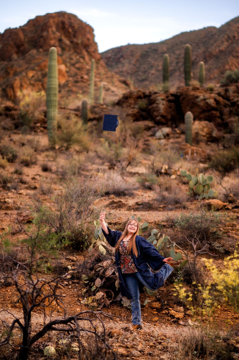 Graduate tosses cap in Arizona desert with saguaro cacti and rocky mountains backdrop.