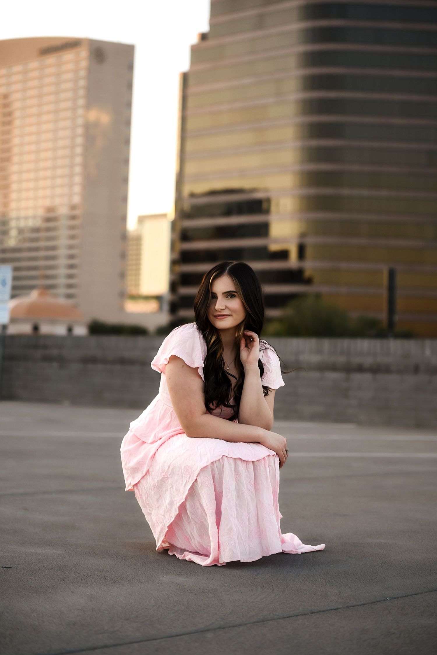 Young woman in pink maxi dress crouching on rooftop with city skyscrapers in background at sunset.