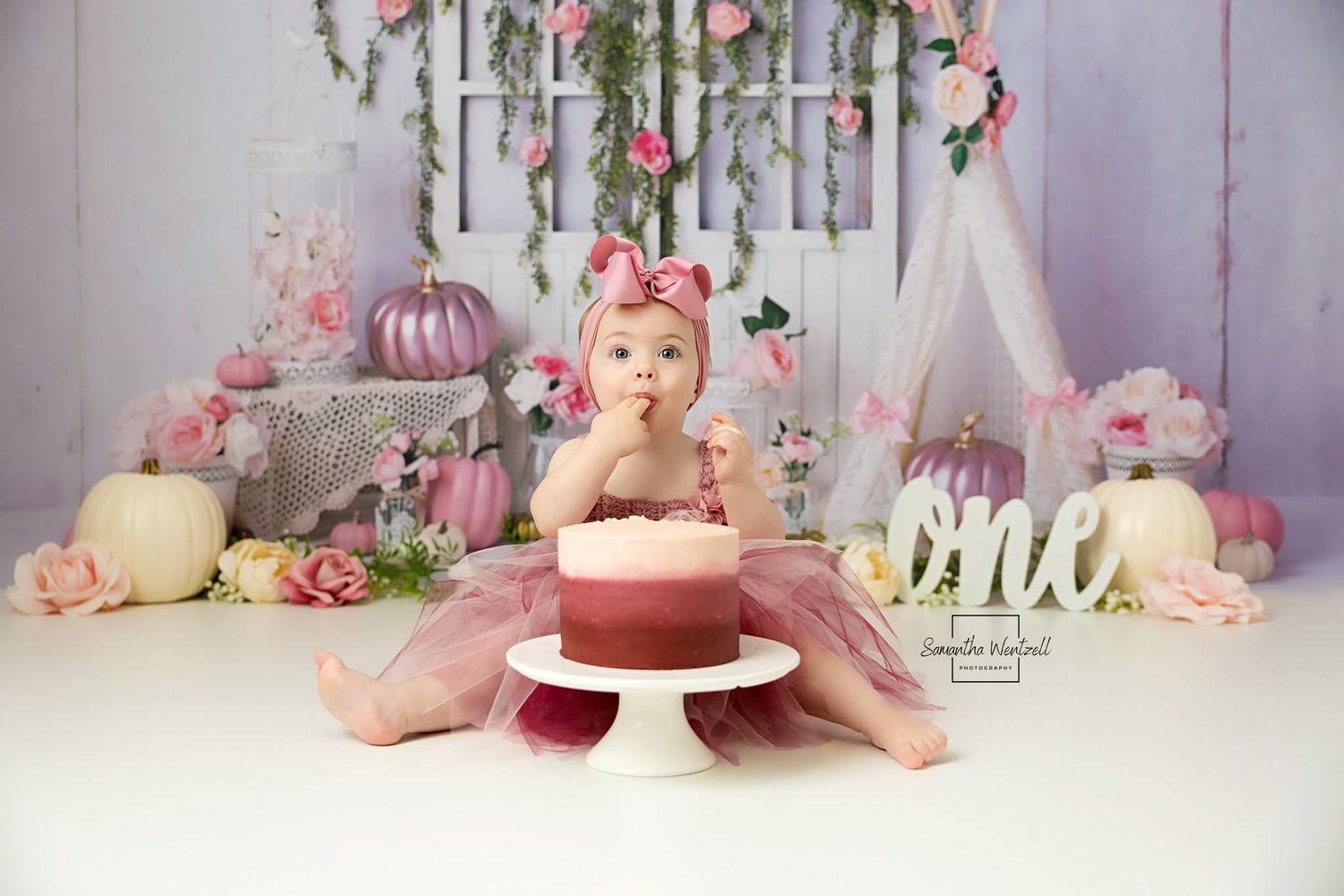 Baby girl in pink tutu sits beside a smash cake surrounded by floral decor for a first birthday photo session.