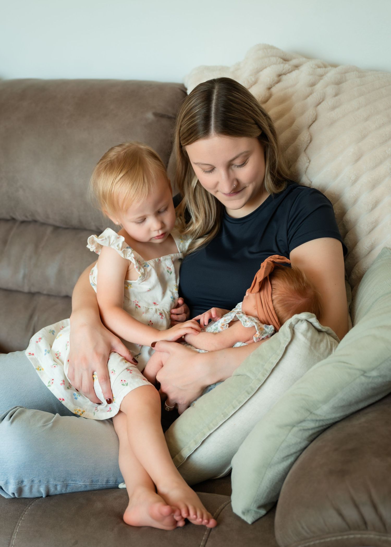 A mother sits on a couch smiling at her newborn baby while her toddler daughter looks on lovingly.