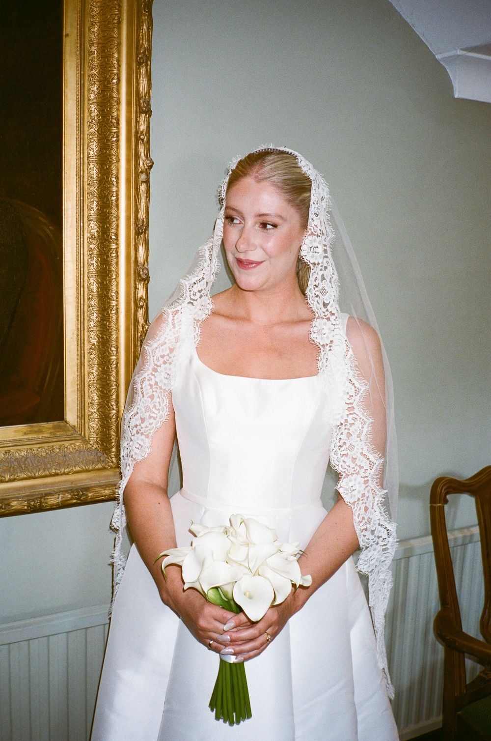 Elegant bride in white gown and lace mantilla veil holding white calla lily bouquet, standing before a gold-framed painting.