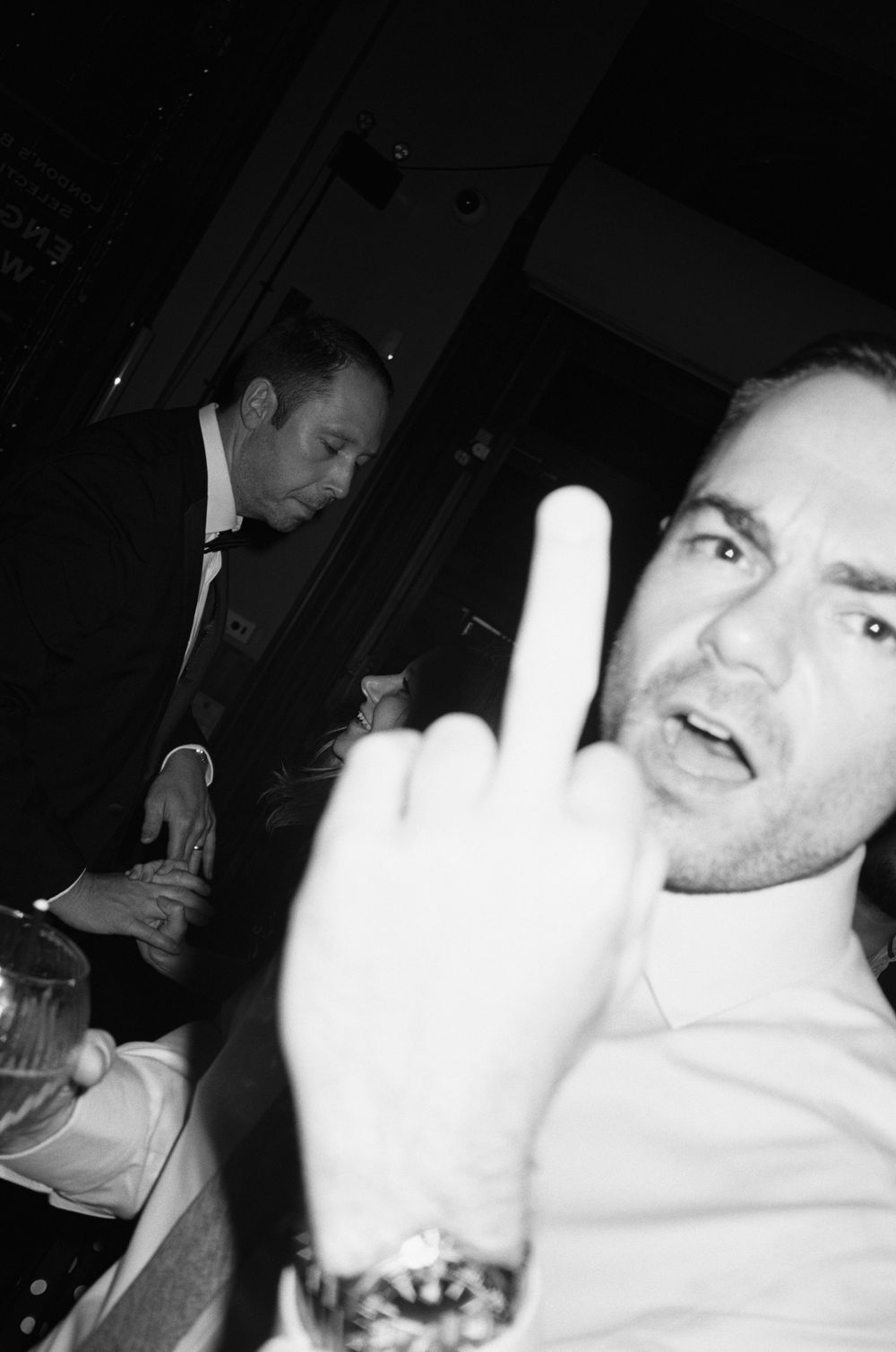 Black and white photo of a man giving the middle finger at a formal event with another man in a tuxedo in the background.