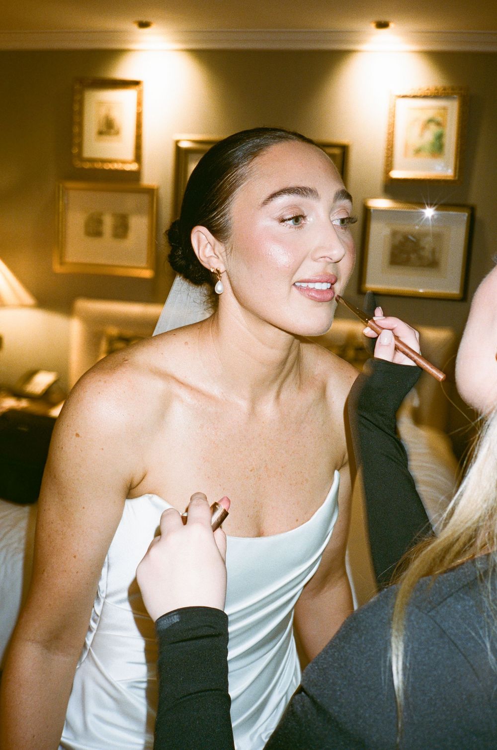 Bride in white strapless gown gets lipstick applied by makeup artist in warmly lit hotel room.