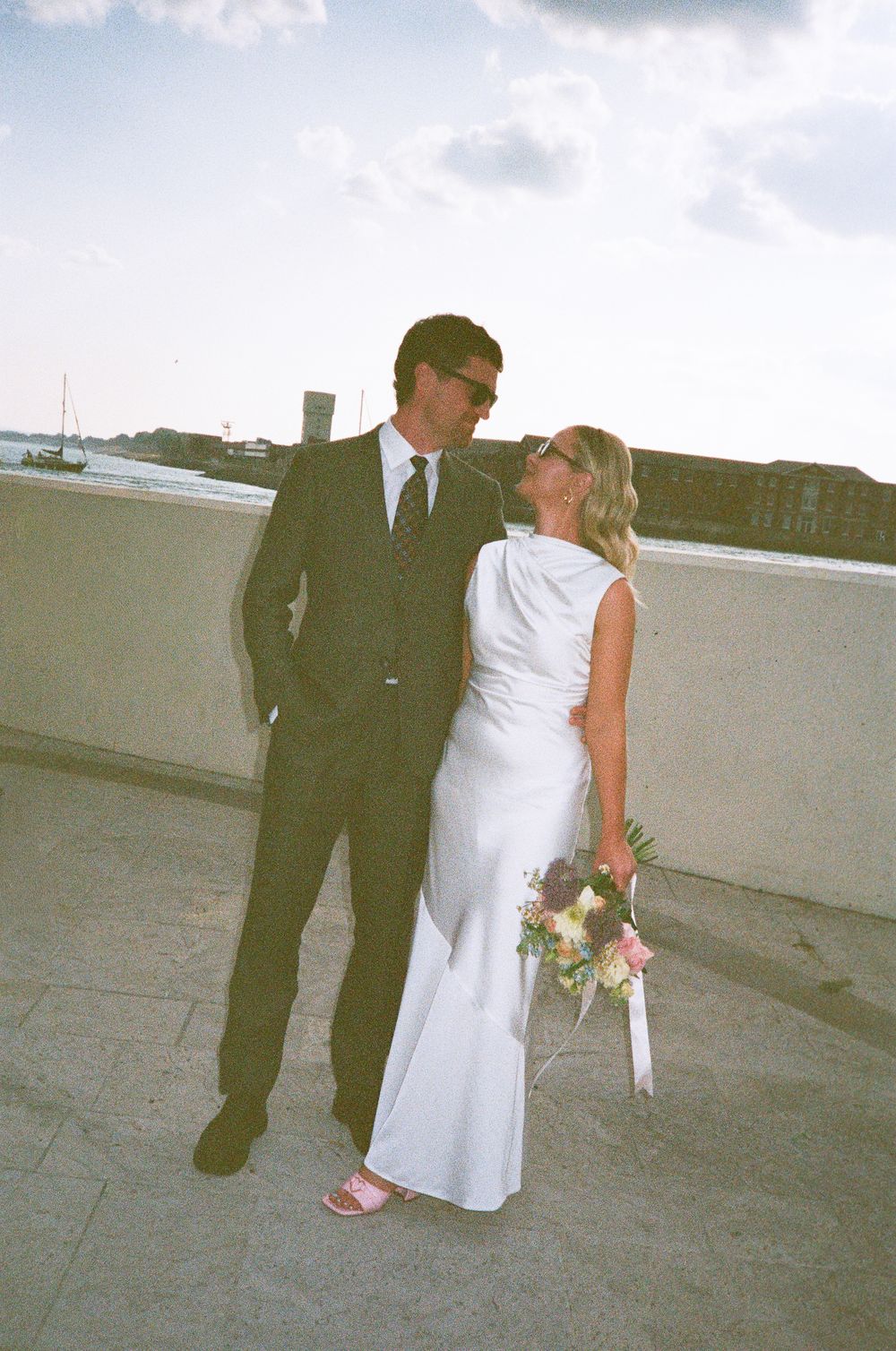 Bride in white gown holding bouquet stands with groom in dark suit on rooftop terrace at outdoor wedding.