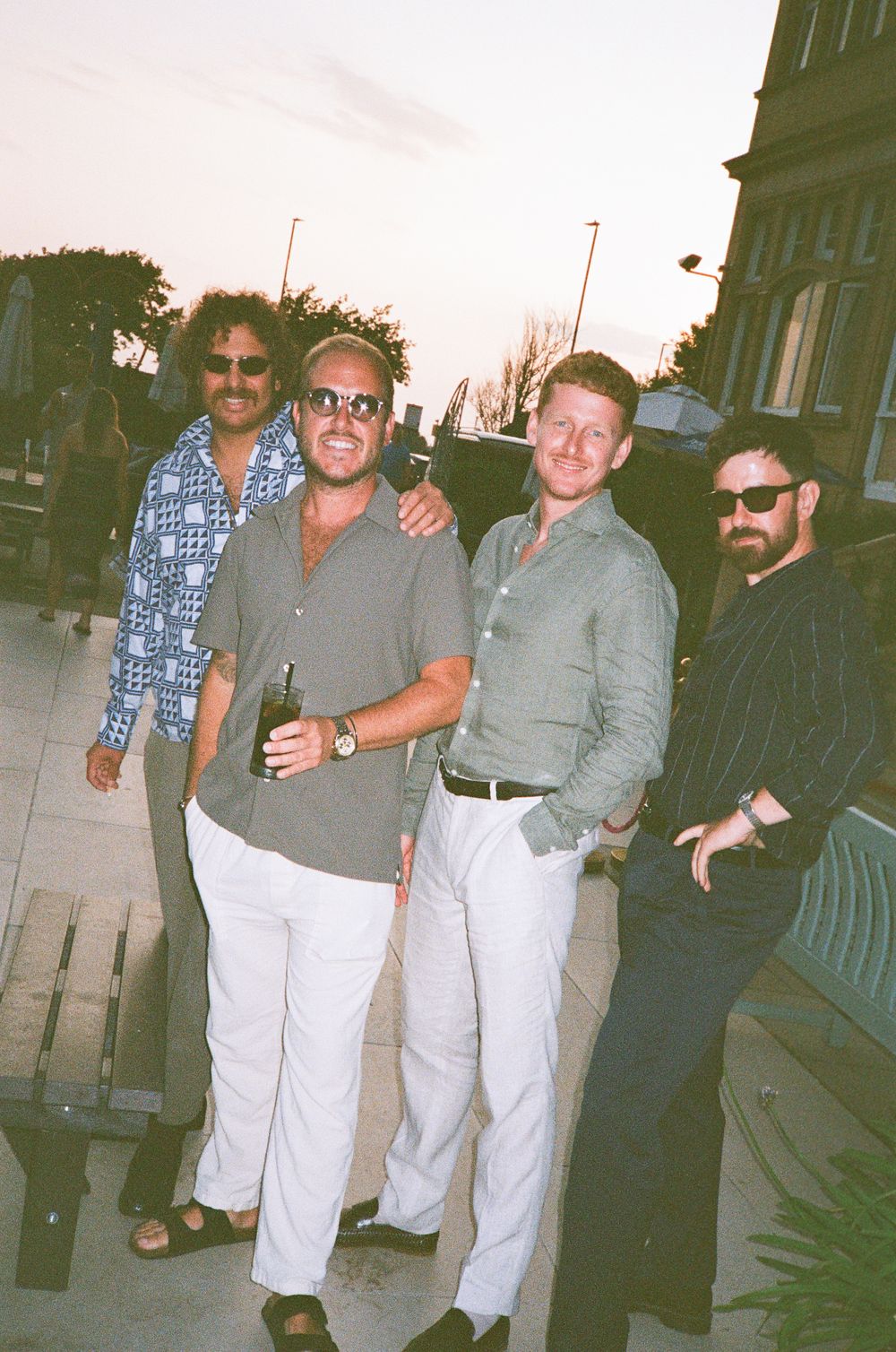 Four men in casual summer attire pose together outdoors at dusk, smiling for a group photo.