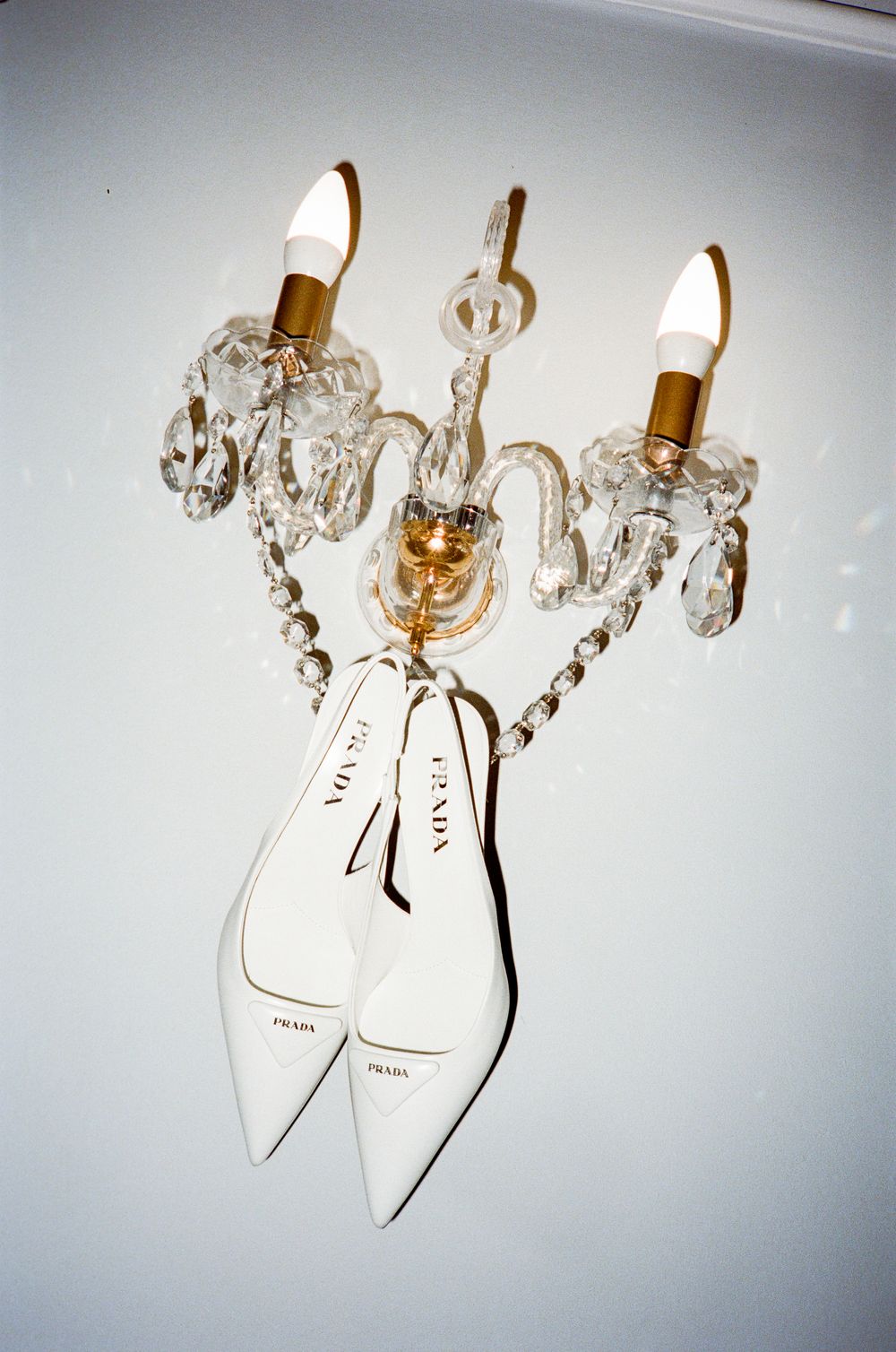White Prada pointed-toe heels hanging from a crystal wall sconce light fixture against a white wall.