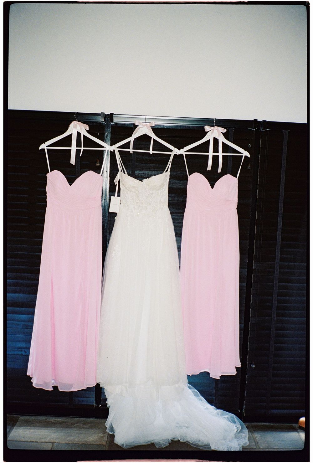 White wedding gown flanked by two pink bridesmaid dresses hanging on white hangers against dark folding doors.