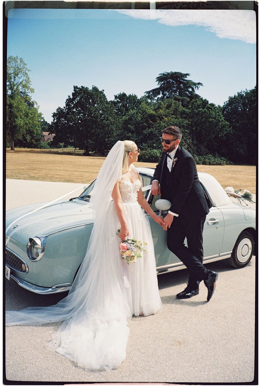 Bride and groom pose beside a vintage pale blue car on their wedding day, surrounded by lush green trees.