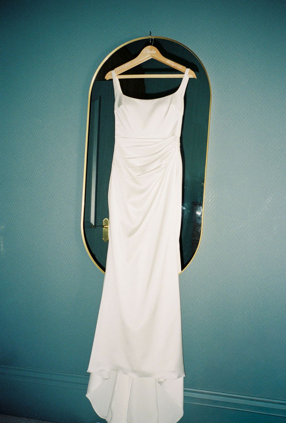 Elegant white sleeveless wedding gown hanging on a wooden hanger against a teal wall with gold mirror.