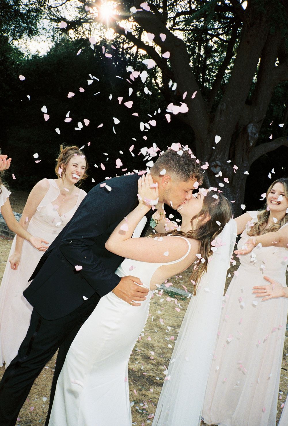 Bride and groom share a kiss surrounded by bridesmaids tossing pink confetti petals outdoors.