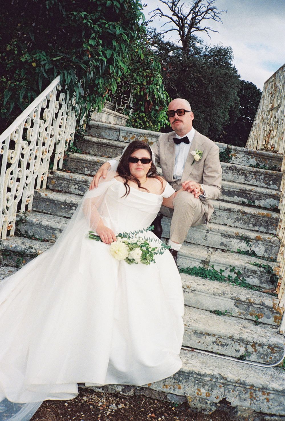 Bride in white gown with bouquet and groom in tan suit with bow tie pose on stone steps outdoors.