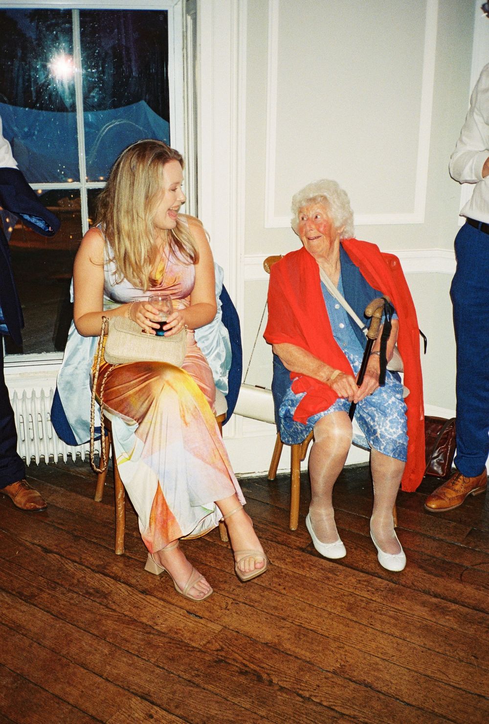 Two women laughing together at a party, one young in colorful dress, one elderly in red shawl holding a cane.