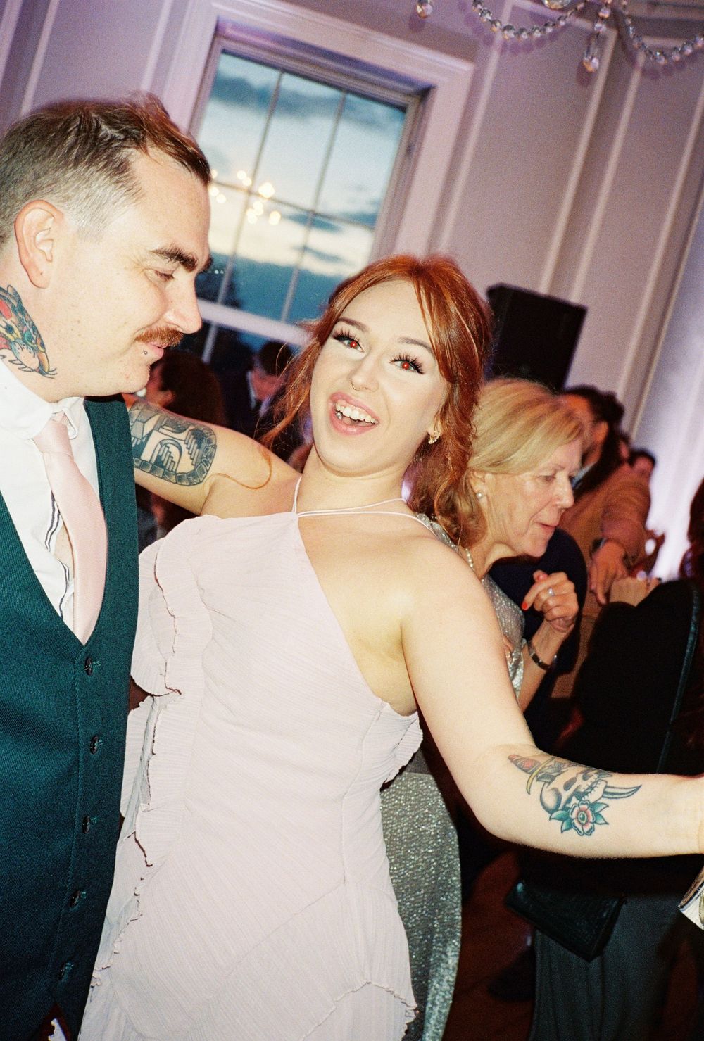 Couple dancing at wedding reception, man with neck tattoos in teal suit, woman in pink dress with floral arm tattoo.