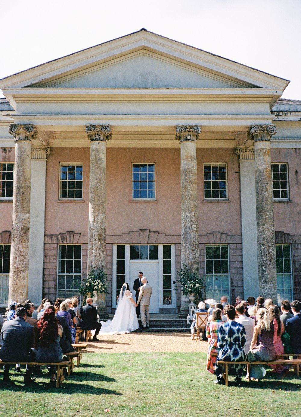 Outdoor wedding ceremony at a grand pink neoclassical mansion with tall columns, guests seated on wooden benches.