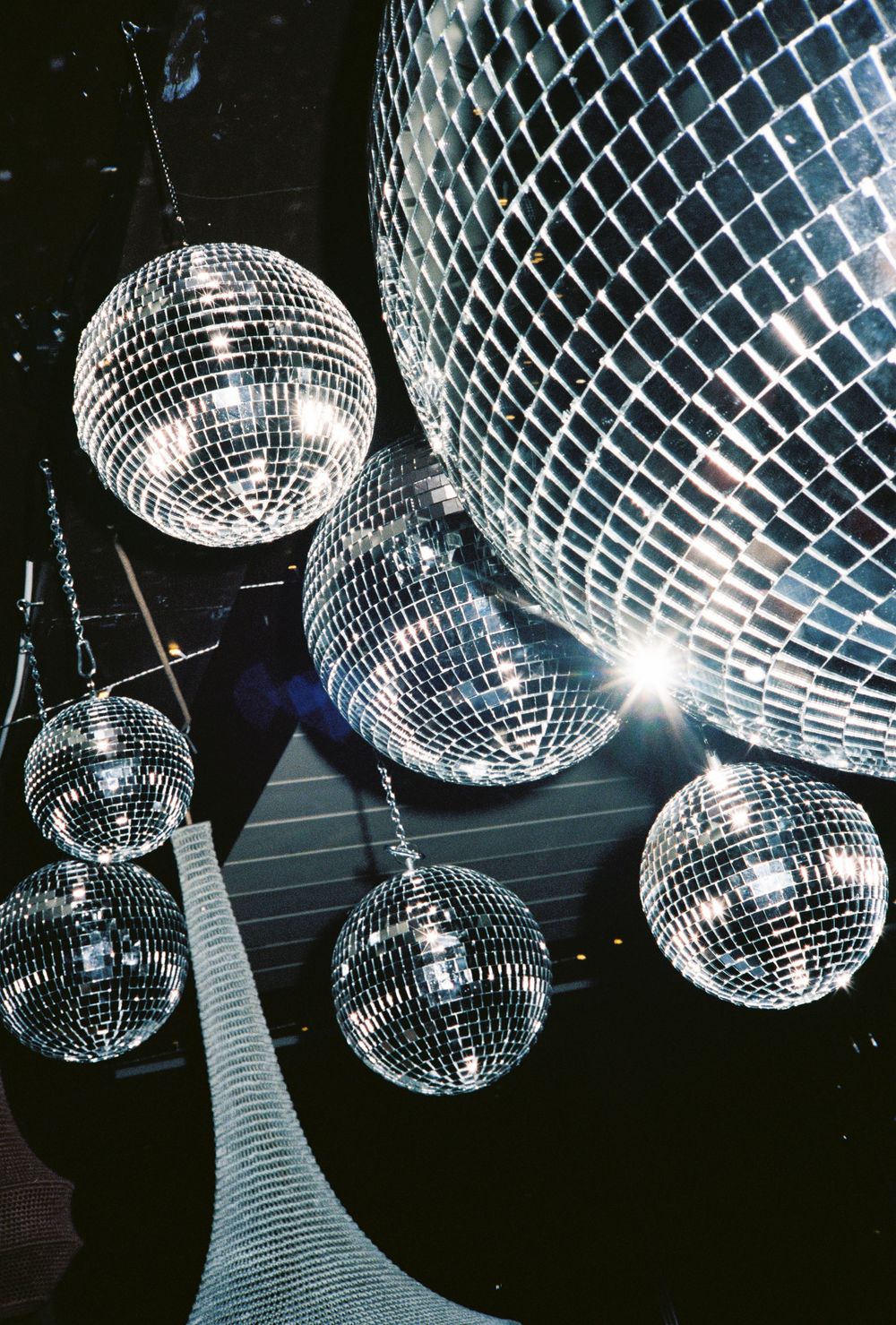 Multiple silver disco mirror balls hanging from a dark ceiling, reflecting sparkling light in a nightclub setting.