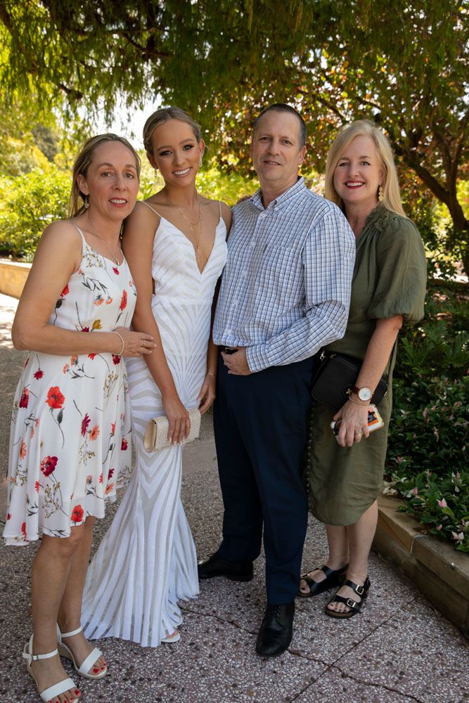 Four people posing outdoors in formal attire, including a woman in a white gown, at a garden event.