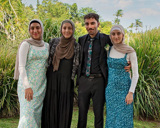Four people posing outdoors at a formal event, three women in hijabs and a man in a black suit, surrounded by lush greenery.