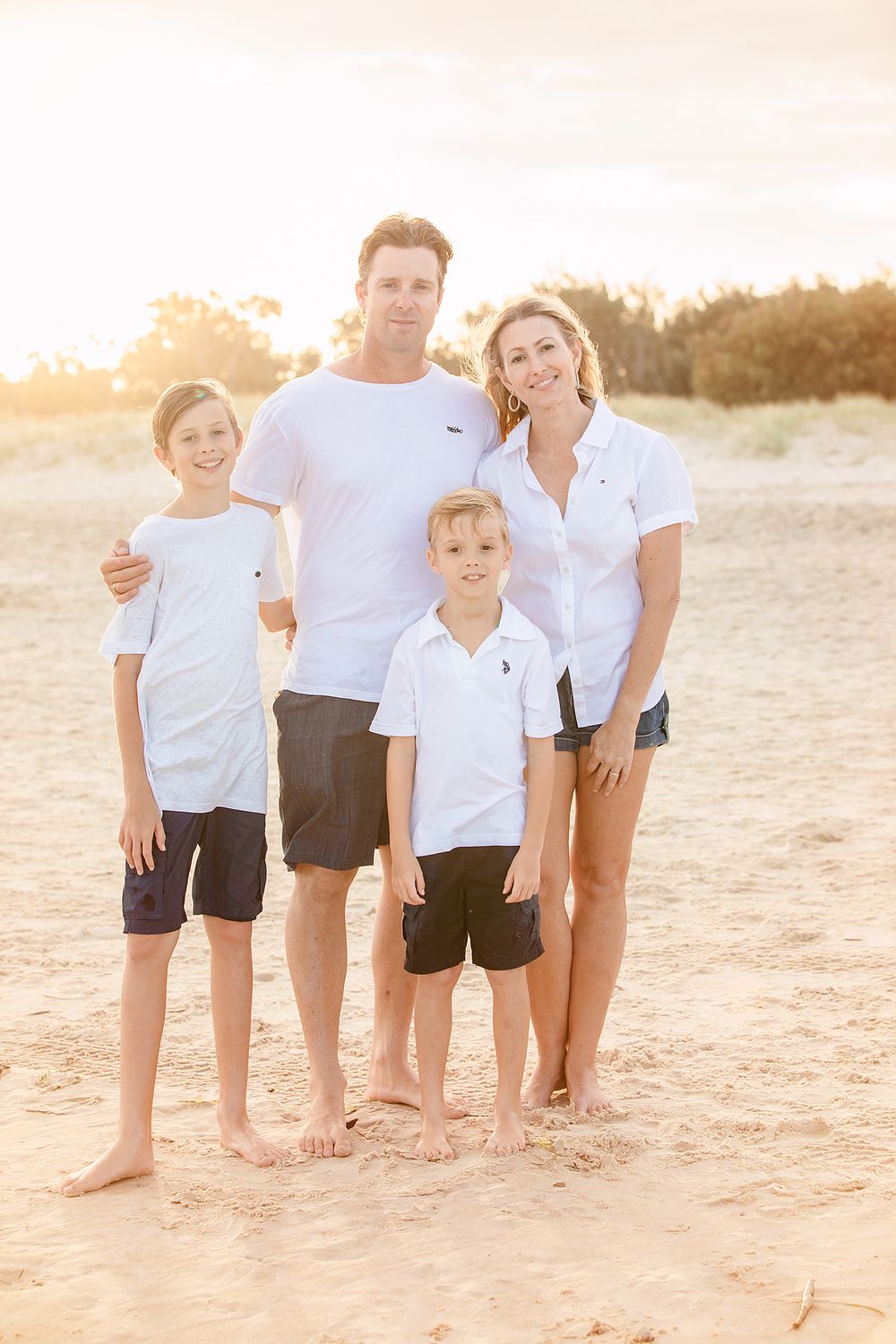 Family of four in white shirts posing barefoot on sandy beach at golden sunset hour.