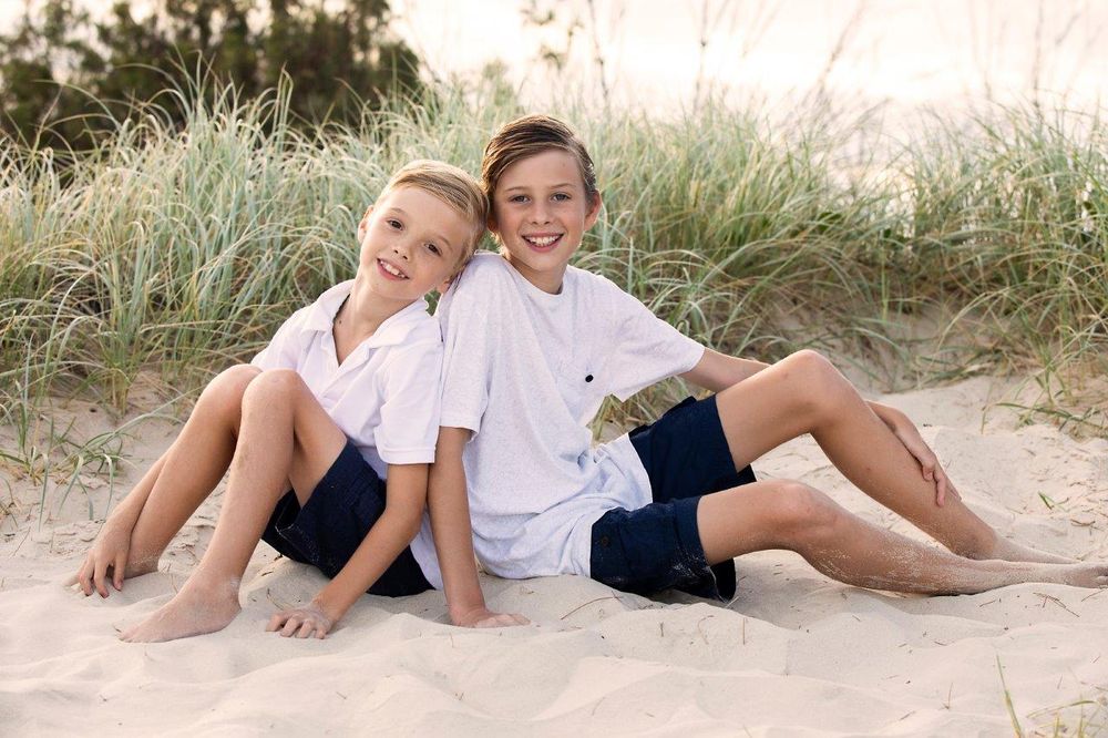 Two smiling boys in white shirts and navy shorts sit back-to-back on sandy beach dunes.