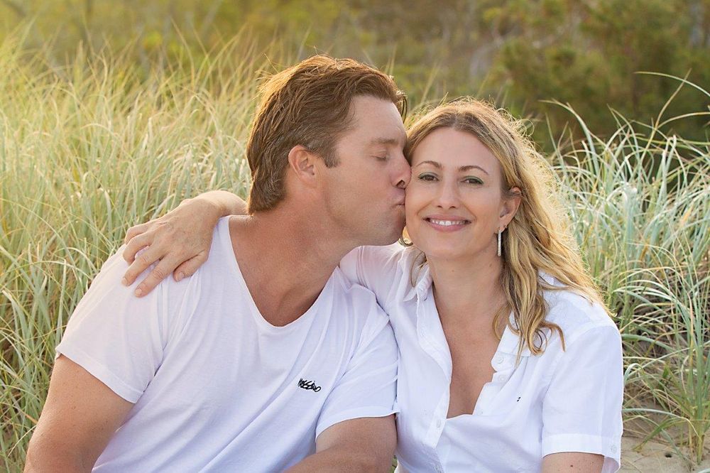 Couple in white shirts sharing a romantic kiss on a sandy beach surrounded by tall grass.