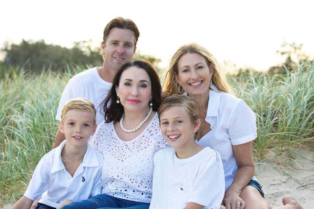 Family of five smiling together on a sandy beach, wearing white outfits, with green dune grass in the background.