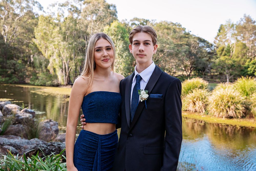 A young couple dressed in formal attire poses outdoors near a pond surrounded by lush greenery.