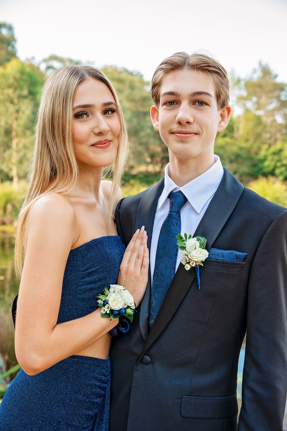 A teenage couple dressed for prom, girl in blue gown with corsage, boy in dark suit with boutonniere, outdoors.
