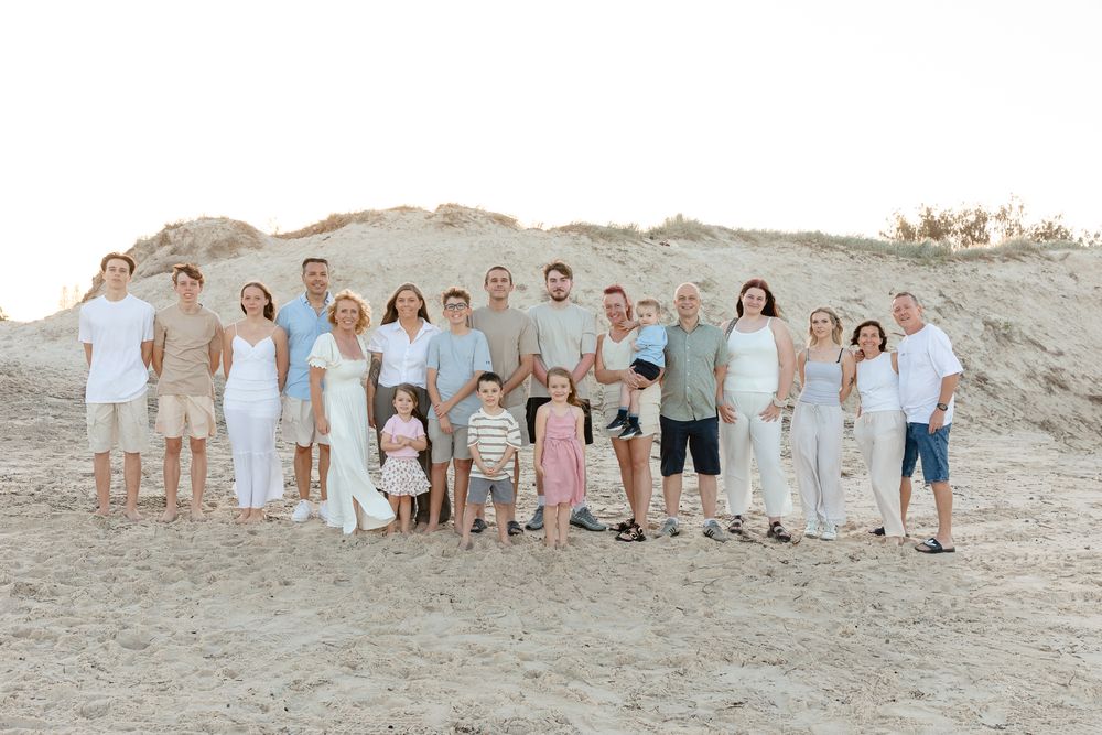 Large family group photo on sandy beach, wearing white and light casual outfits, posing together with sand dunes in background.