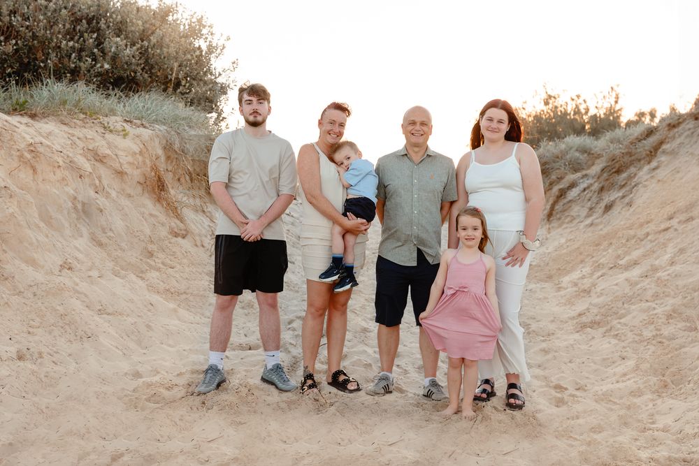 Family of six posing together on sandy beach dunes at golden hour, smiling for a portrait photo.