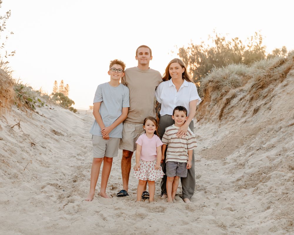 Family of five posing on sandy beach dunes at golden hour, two adults and three children smiling outdoors.