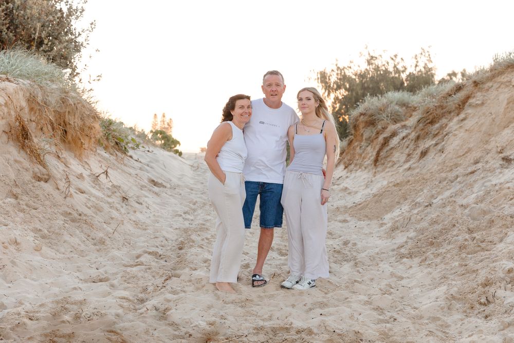 Family of three smiling together on sandy beach dunes at golden hour, wearing casual white and blue outfits.