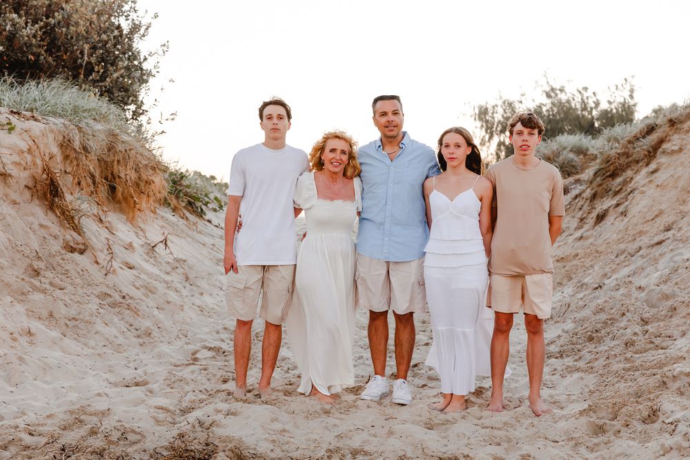 Family of five posing together on a sandy beach path at golden hour, wearing coordinated neutral outfits.