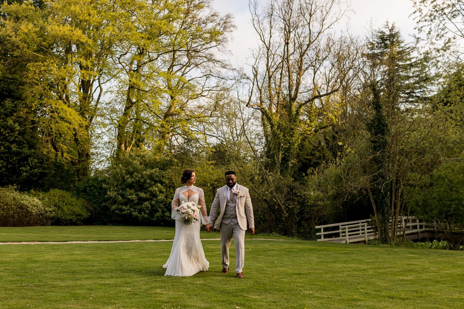 Bride and groom share a romantic kiss on a wooden bridge surrounded by lush green trees in a garden setting.