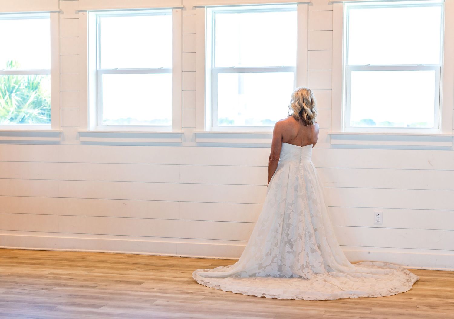Bride in elegant lace wedding gown with train stands facing windows in bright white shiplap venue.