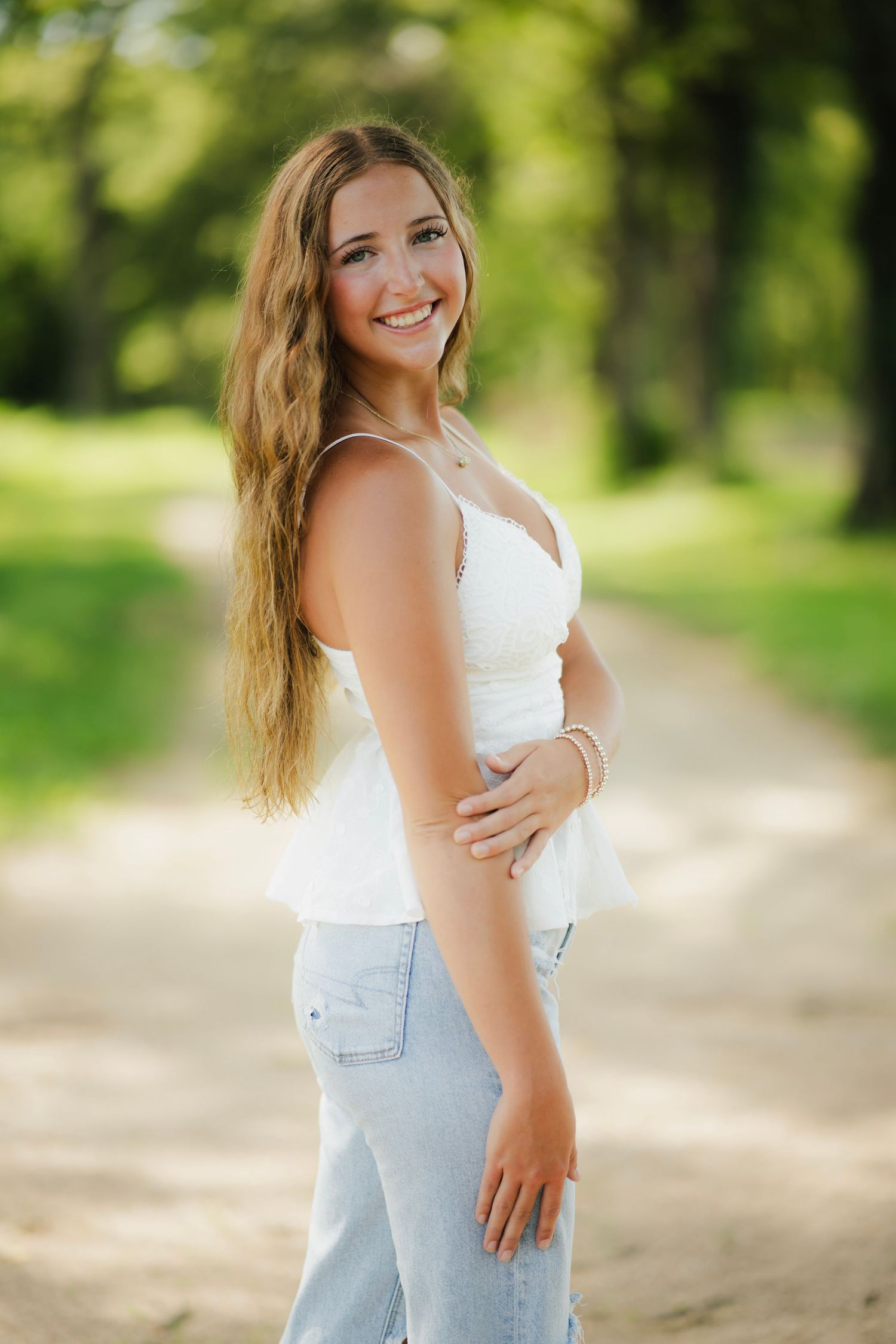 Young woman in white top and ripped jeans walks barefoot down a tree-lined dirt path in a lush green park.