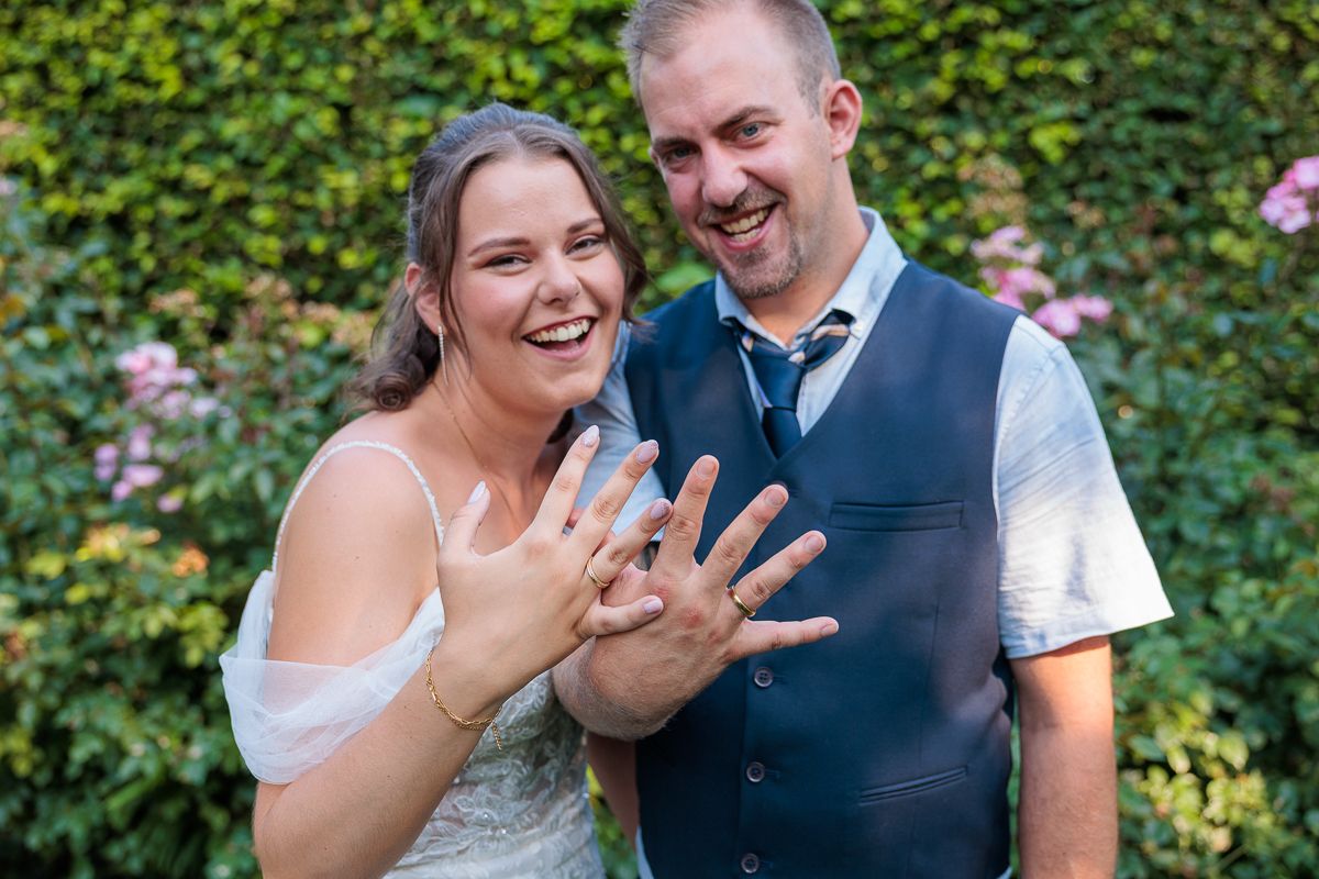 A bride and groom pose together in a lush green outdoor setting, holding a dried floral bouquet on their wedding day.